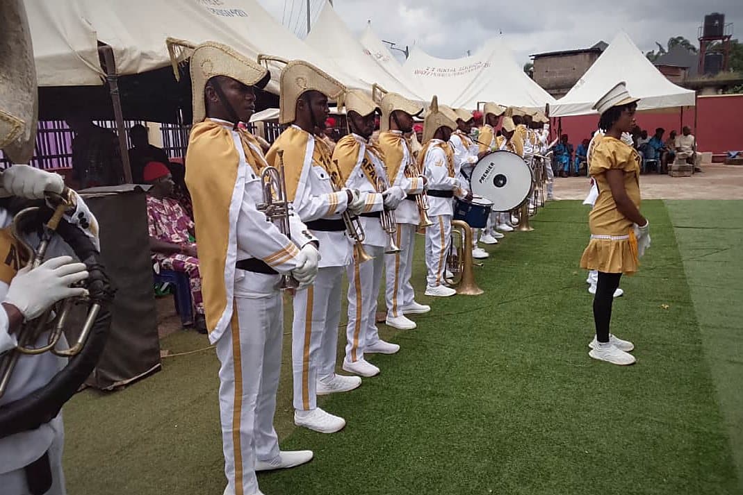 A brass band drawn up in white and gold uniform, instruments raised, before a service