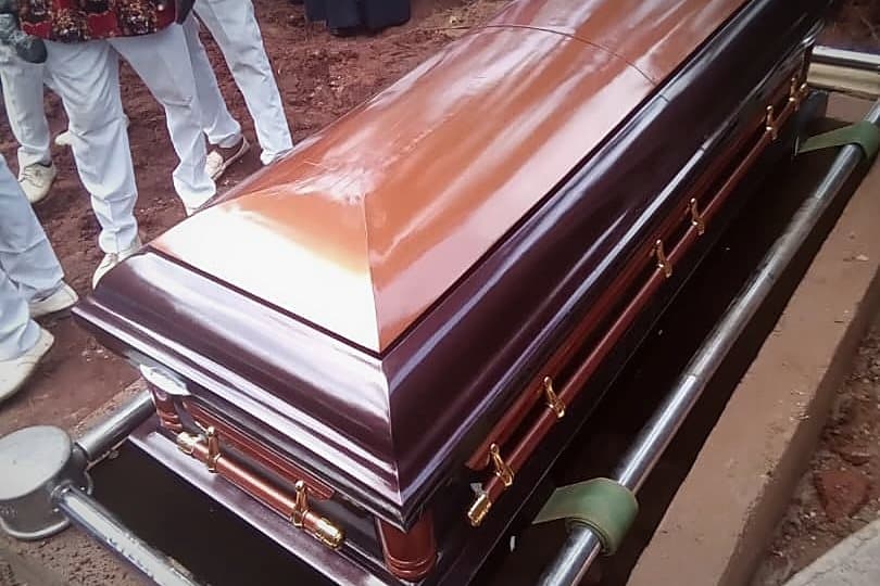 A mahogany casket at the graveside on its lowering frame, clergy standing beyond it