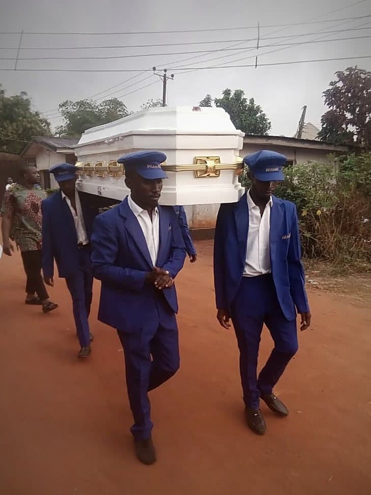 Four pallbearers in blue suits and caps carrying a white casket along a red-earth road